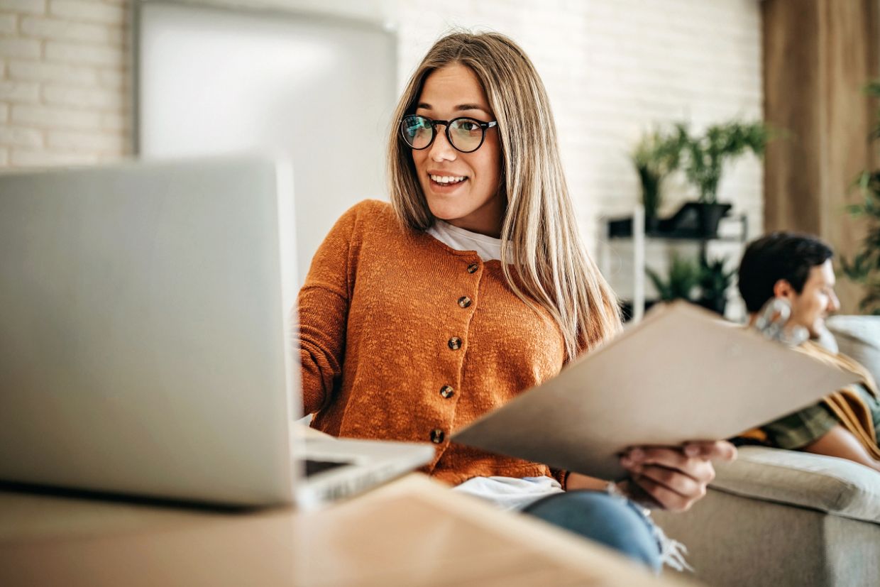 Frau am Tisch, die lächelnd an ihrem Laptop arbeitet. Im Hintergrund sitzt ein Mann auf der Couch.