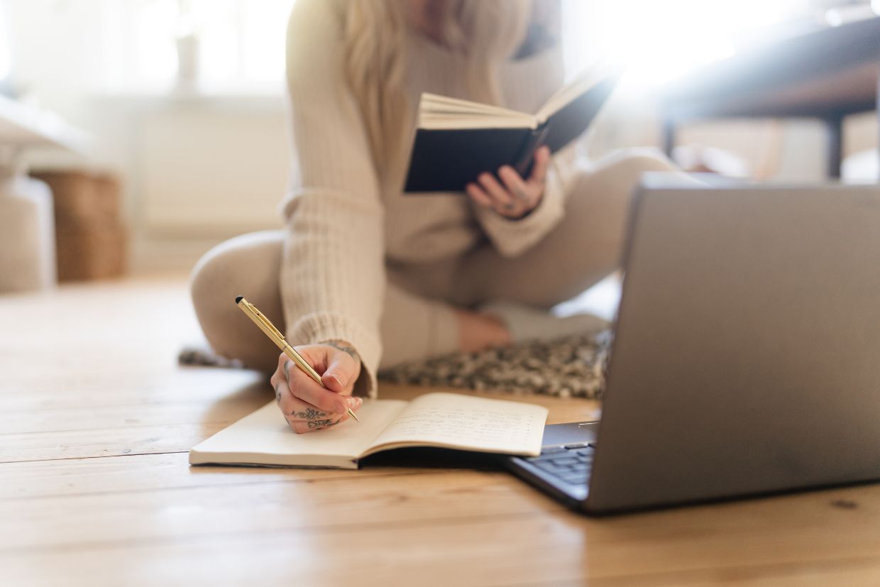 Ein Bild von einer Frau am Laptop, die auf dem Boden sitzt und mit einem Notizbuch arbeitet.